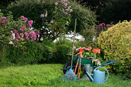 The image depicts a well-maintained garden area with a lush, green lawn surrounded by colorful flowering shrubs and bushes, including pink and purple blossoms. In the foreground, various garden tools and accessories are visible, such as a blueish-green watering can, a garden rake, and a gardening bucket, suggesting ongoing maintenance activities. Behind the tools, a small, neat flower bed borders a gravel or stone path that separates the lawn from a darker, shaded area with taller trees and dense foliage in the background. The garden appears to be in rural or suburban Dulwich, with natural sunlight illuminating the scene, indicating fair weather. The overall scene reflects a tidy, vibrant outdoor space suitable for gardening and lawn care services, with a mixture of natural tones and structured garden elements, illustrating an inviting and well-kept garden environment.