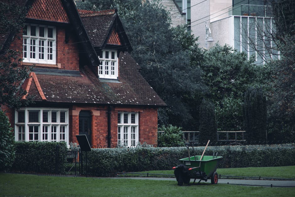 A red brick house with a steep, tiled roof and white-framed windows surrounded by a well-maintained garden. The garden features a neatly trimmed hedge, lush green lawn, and mature trees with dense fol
