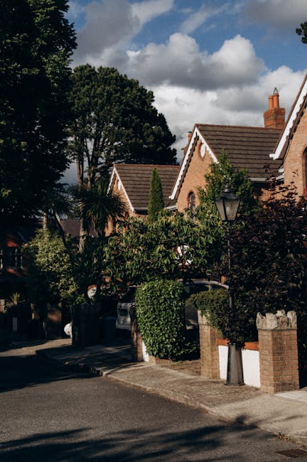 A red brick house with a steep, tiled roof and white-framed windows surrounded by a well-maintained garden. The garden features a neatly trimmed hedge, lush green lawn, and mature trees with dense fol